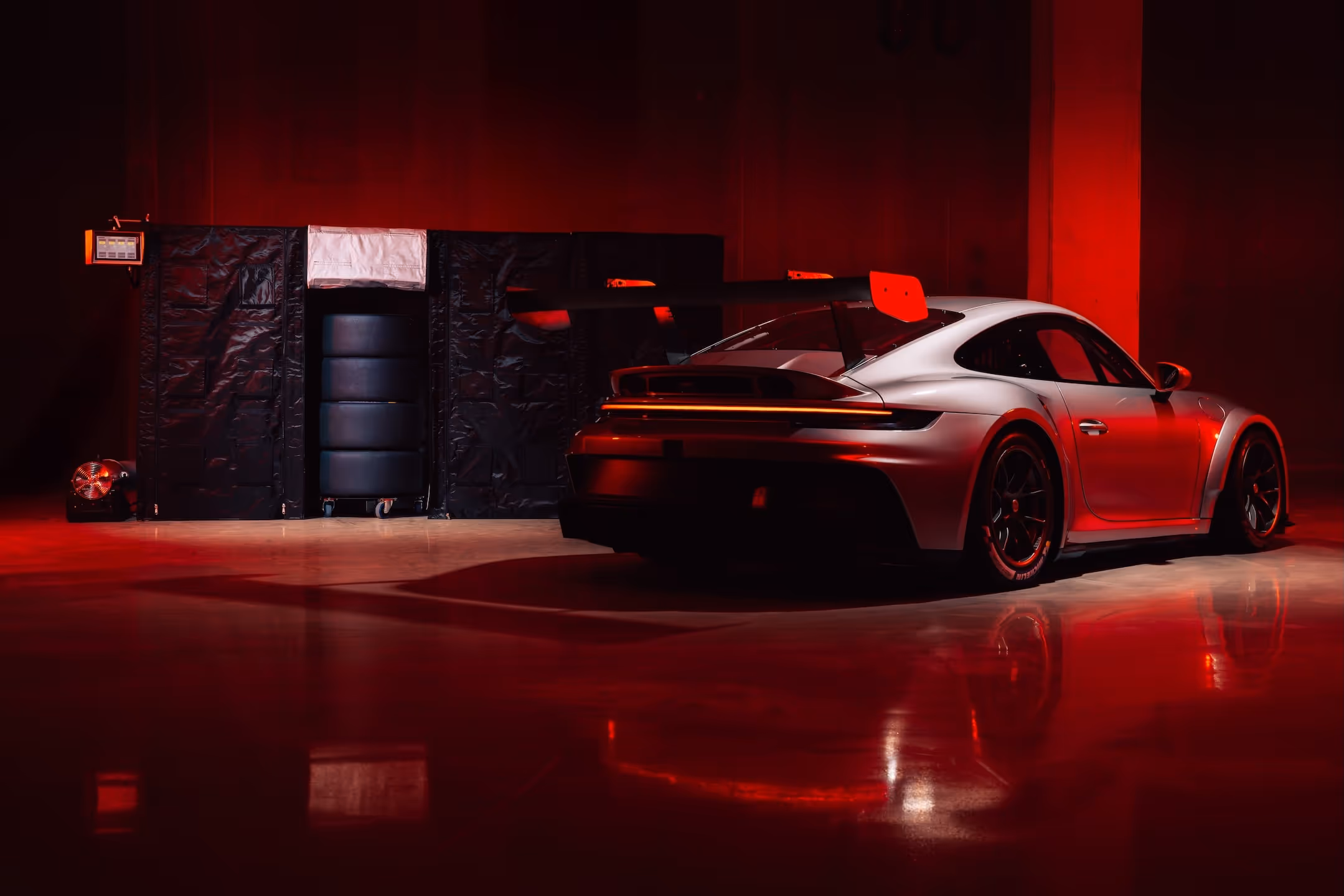 Silver sports car with large rear wing illuminated by red lighting in a dark garage.