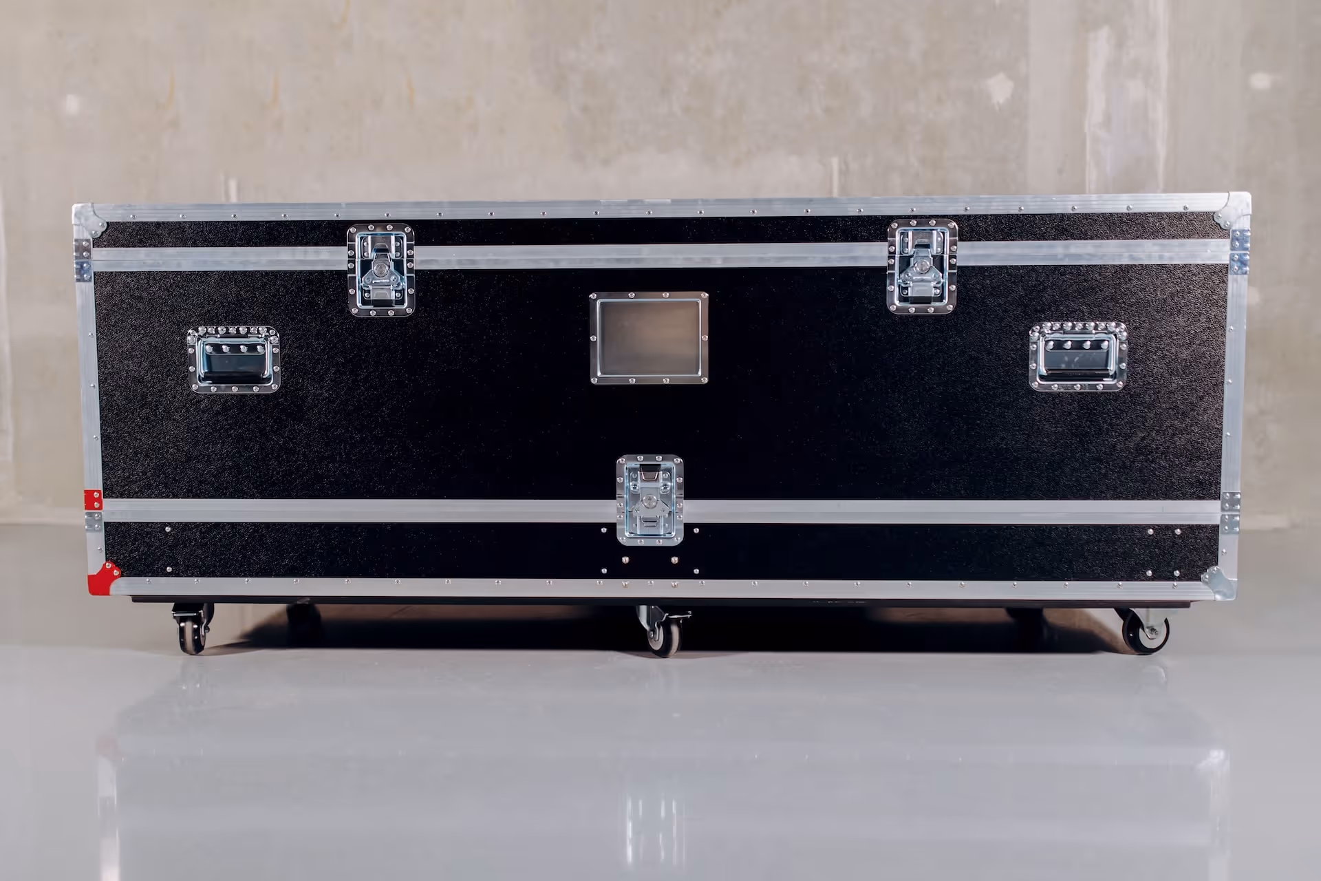 Large black flight case with metal edges and latches on caster wheels against a plain wall.