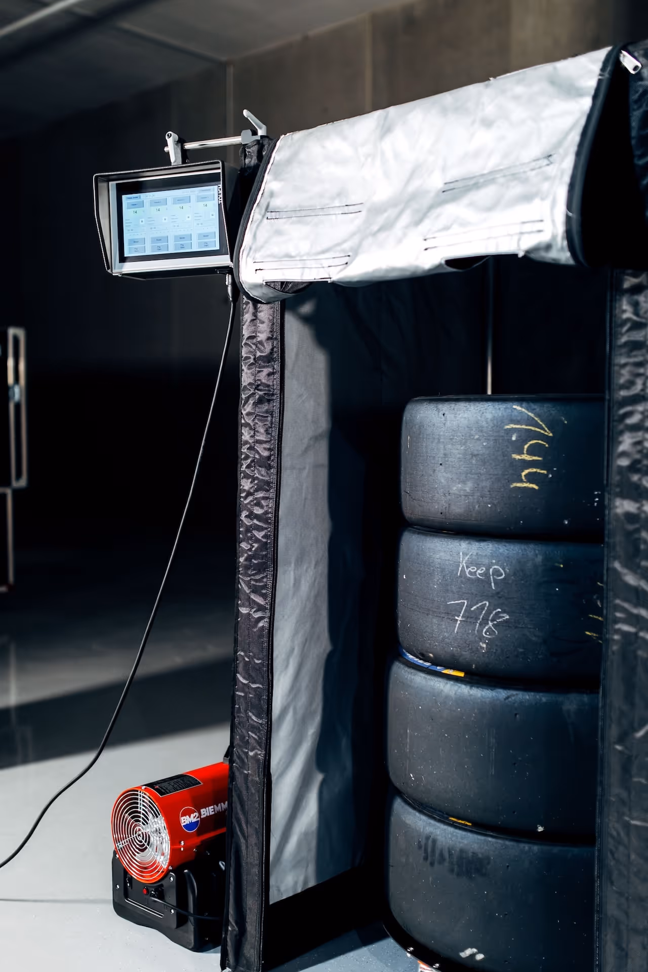 Stack of four black racing tires inside a heated tent with a control screen and a red blower fan outside.