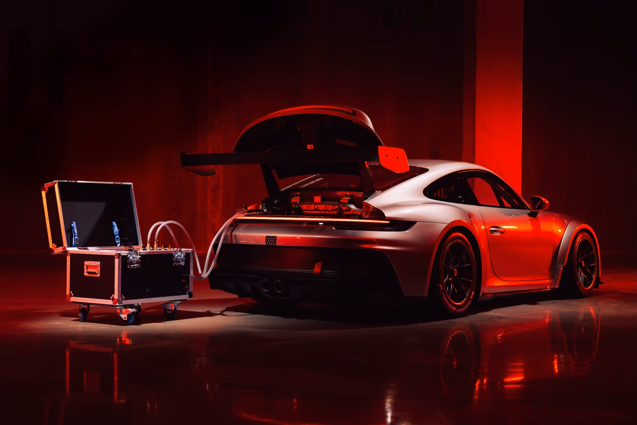 Silver sports car with rear hatch open connected by tubes to a black equipment box in a red-lit room.