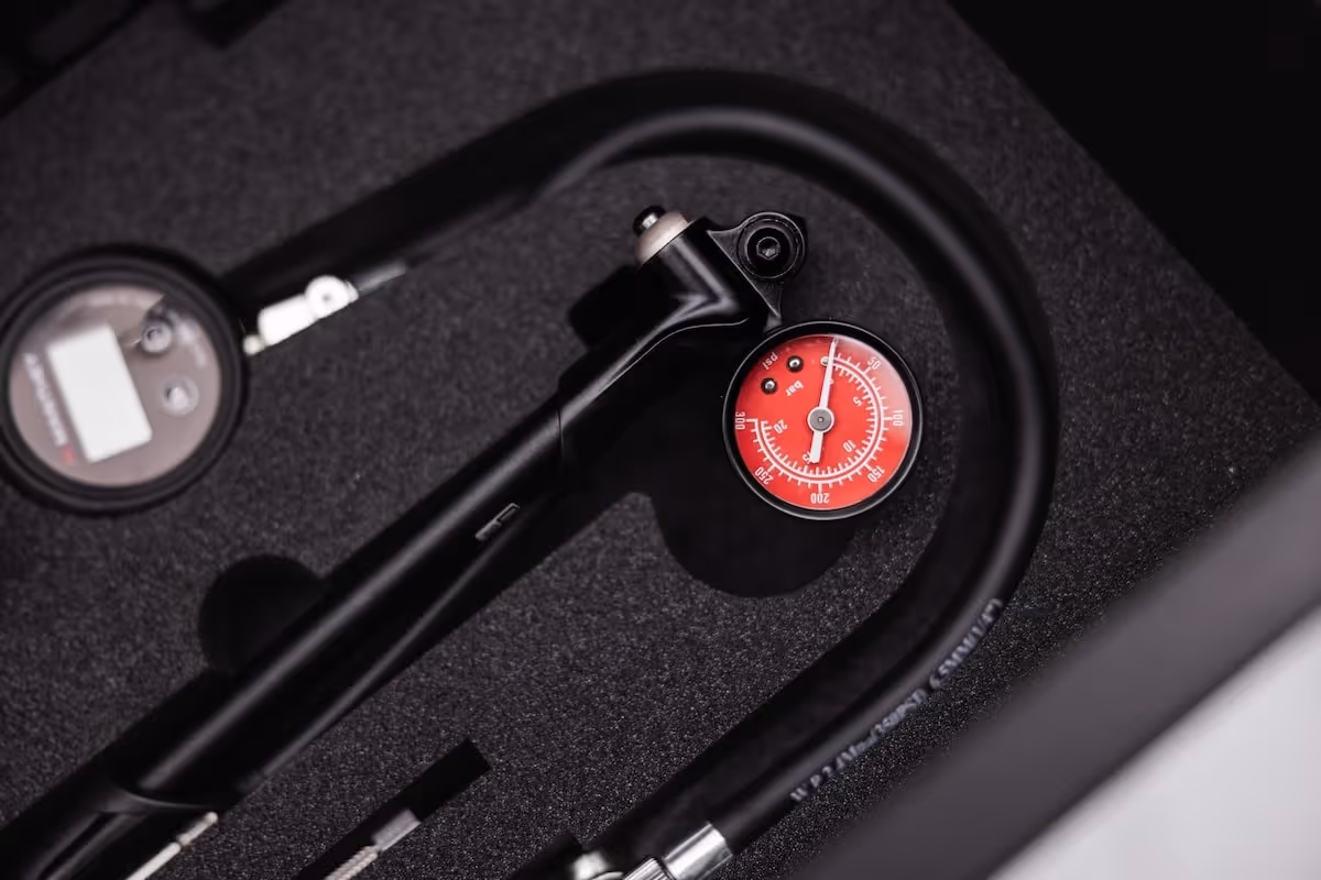 Black tool with a red pressure gauge and a flexible hose, placed on dark foam padding.