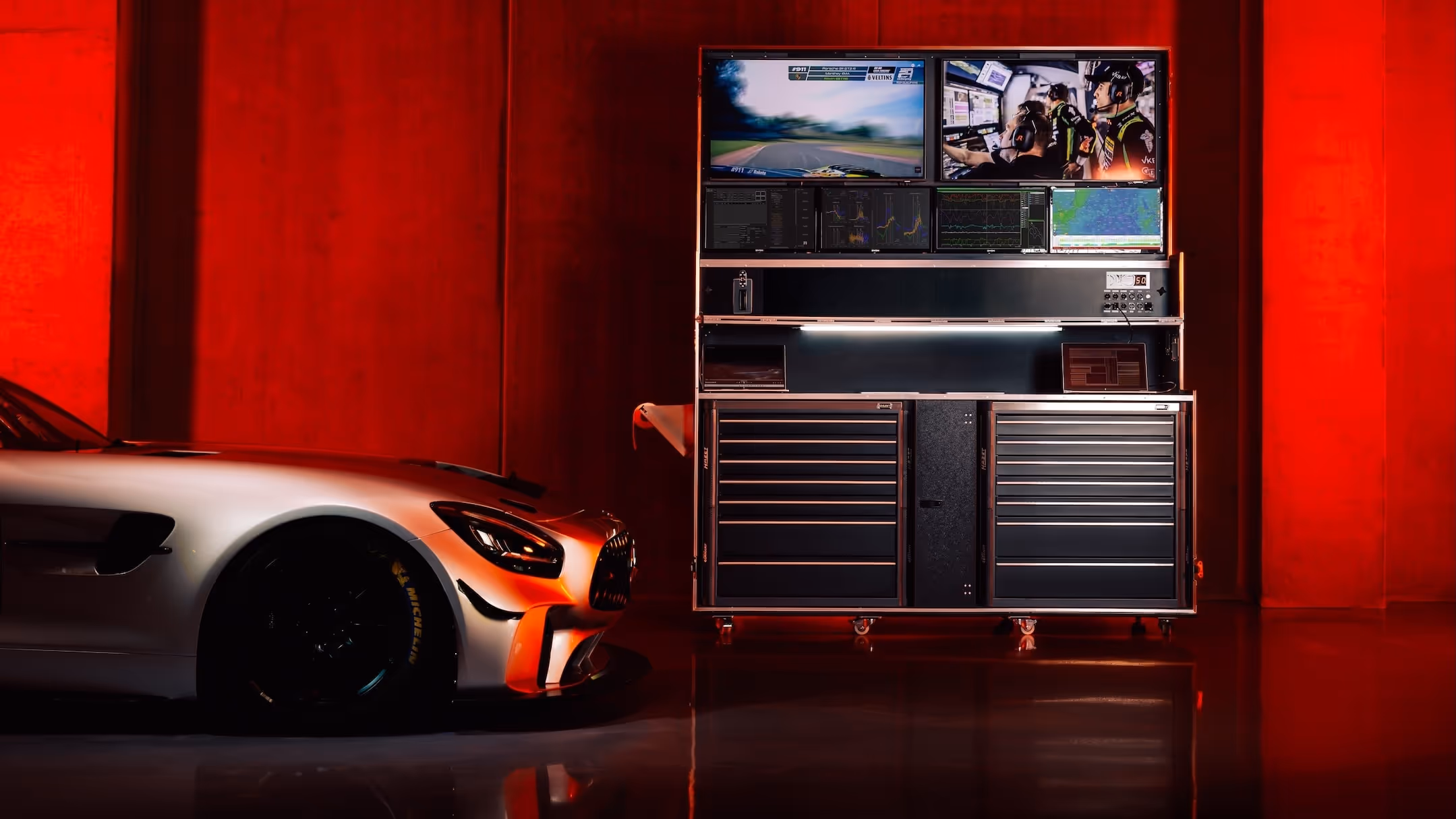 Silver sports car facing a large multi-screen racing telemetry and monitoring system in a red-lit room.