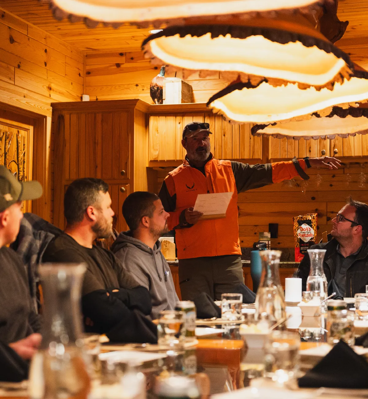 Man in an orange vest holding a paper and speaking to a group seated around a wooden table in a cozy wood-paneled room.