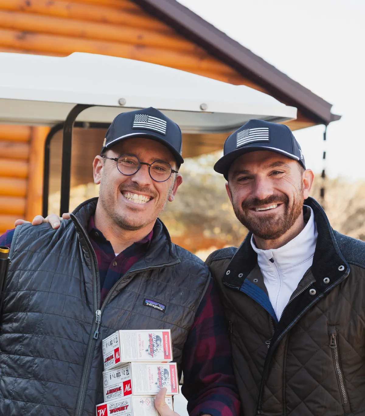 Two men wearing black caps with American flags smiling and posing, one holding ammunition boxes.