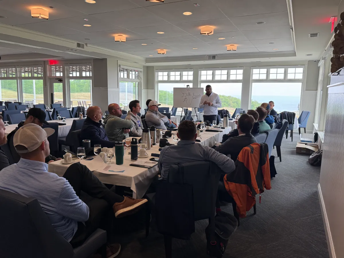 Group of people seated around a conference table attentively listening to a man giving a presentation in a bright room with large windows and a whiteboard.