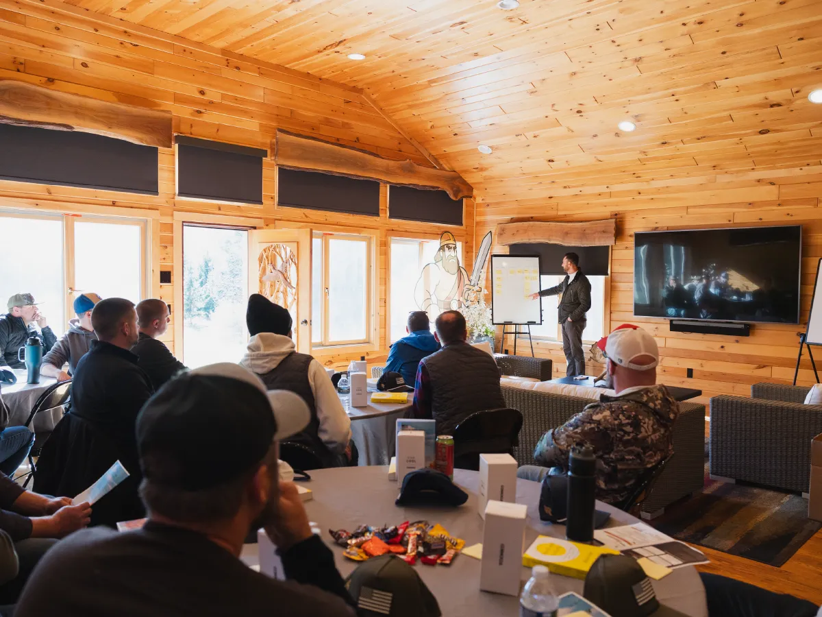 Group of people sitting at round tables attentively listening to a man presenting on a flip chart in a wood-paneled room.