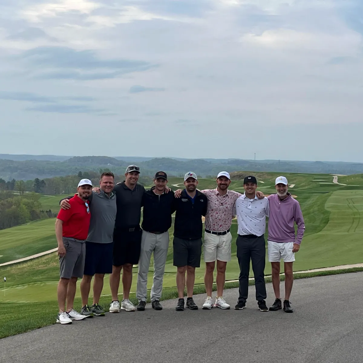 Eight men standing arm in arm on a golf course with rolling hills and cloudy sky in the background.