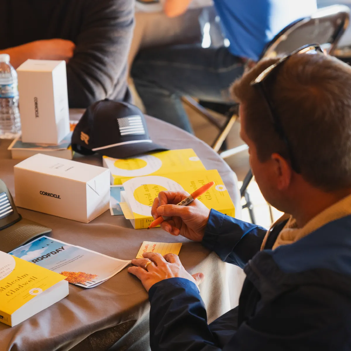 Man wearing sunglasses on his head writing on a yellow sticky note at a table with books, a black cap, and a water bottle.