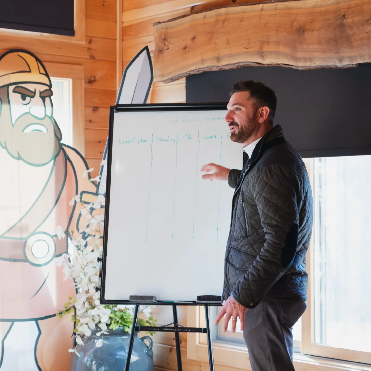 Man in a quilted jacket stands next to a whiteboard with columns, giving a presentation indoors near a large cartoon figure on the wall.