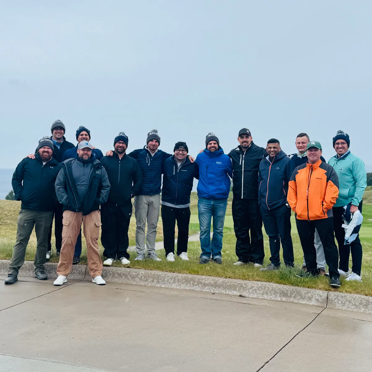 Group of thirteen men standing outdoors on a cloudy day, dressed in jackets, hats, and casual pants on a golf course.