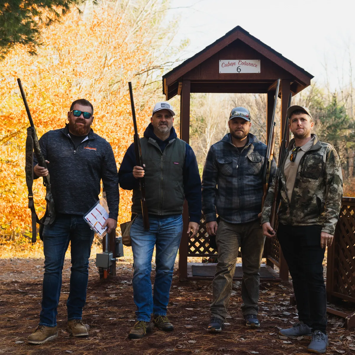 Four men holding shotguns standing outdoors near a wooden shooting station with fall foliage in the background.