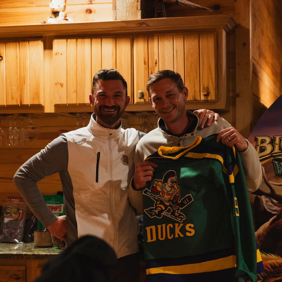 Two men smiling indoors, one holding a green hockey jersey with a duck mascot and the word 'DUCKS'.