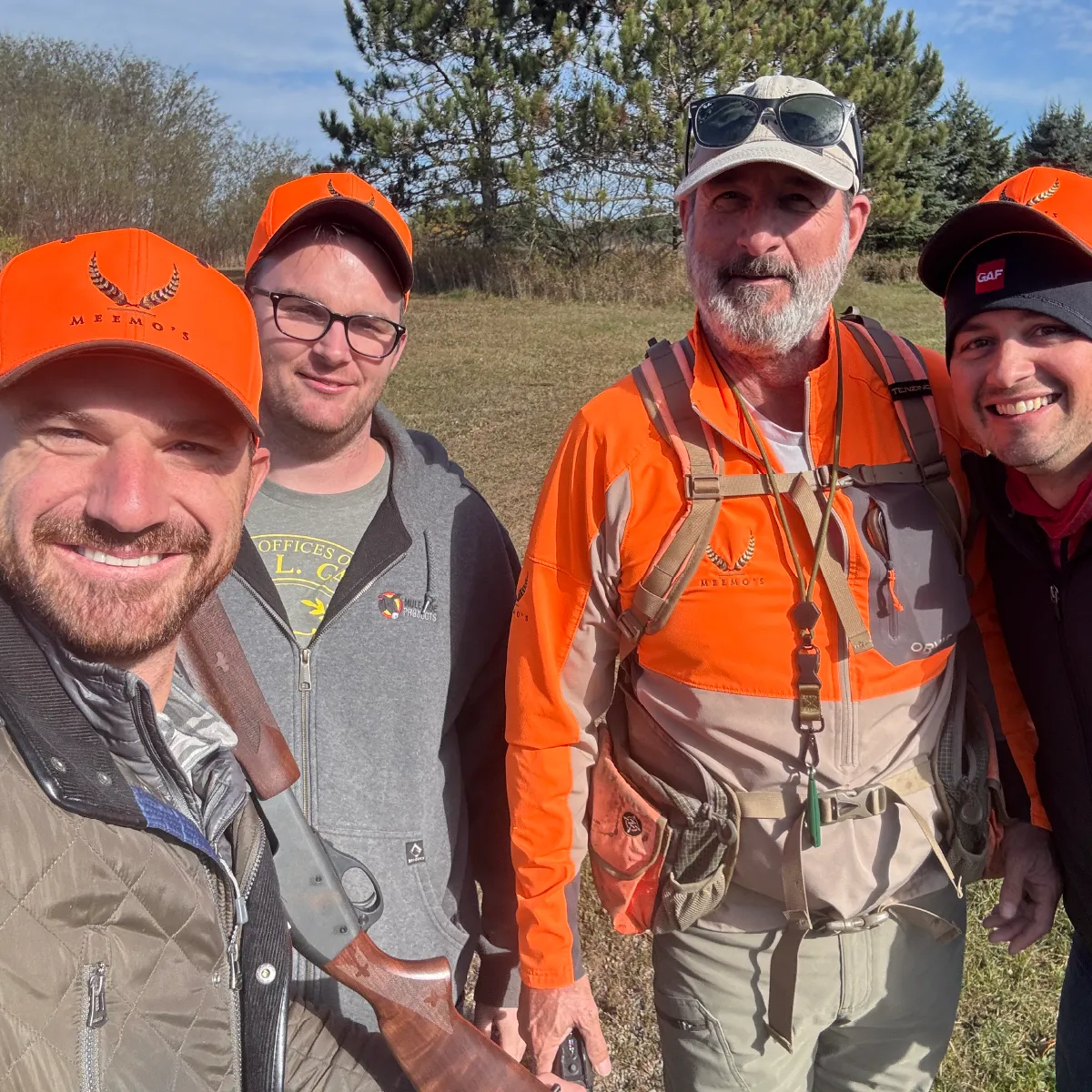 Four men smiling outdoors wearing orange hunting hats and jackets, with one holding a shotgun.