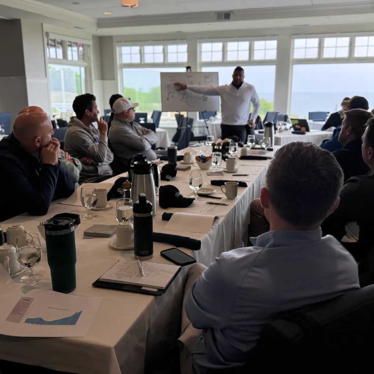 Group of people seated around a table attentively listening to a man presenting with a whiteboard in a bright room with large windows.