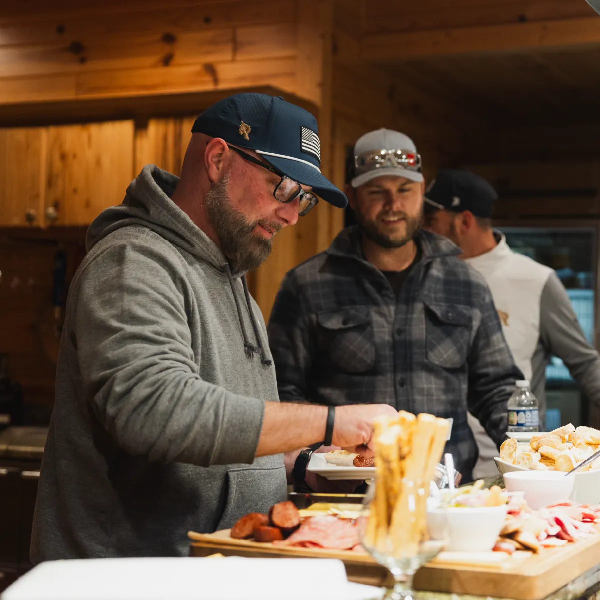 Three men standing at a wooden kitchen counter with a charcuterie board, one man in a gray hoodie and cap spreading food on a plate.