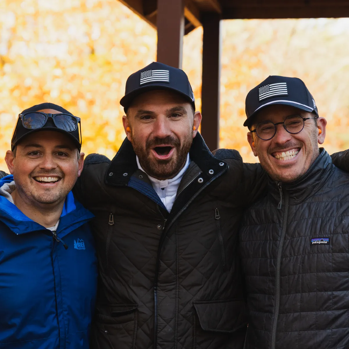Three men wearing outdoor jackets and caps with American flags, smiling and posing closely together outdoors.