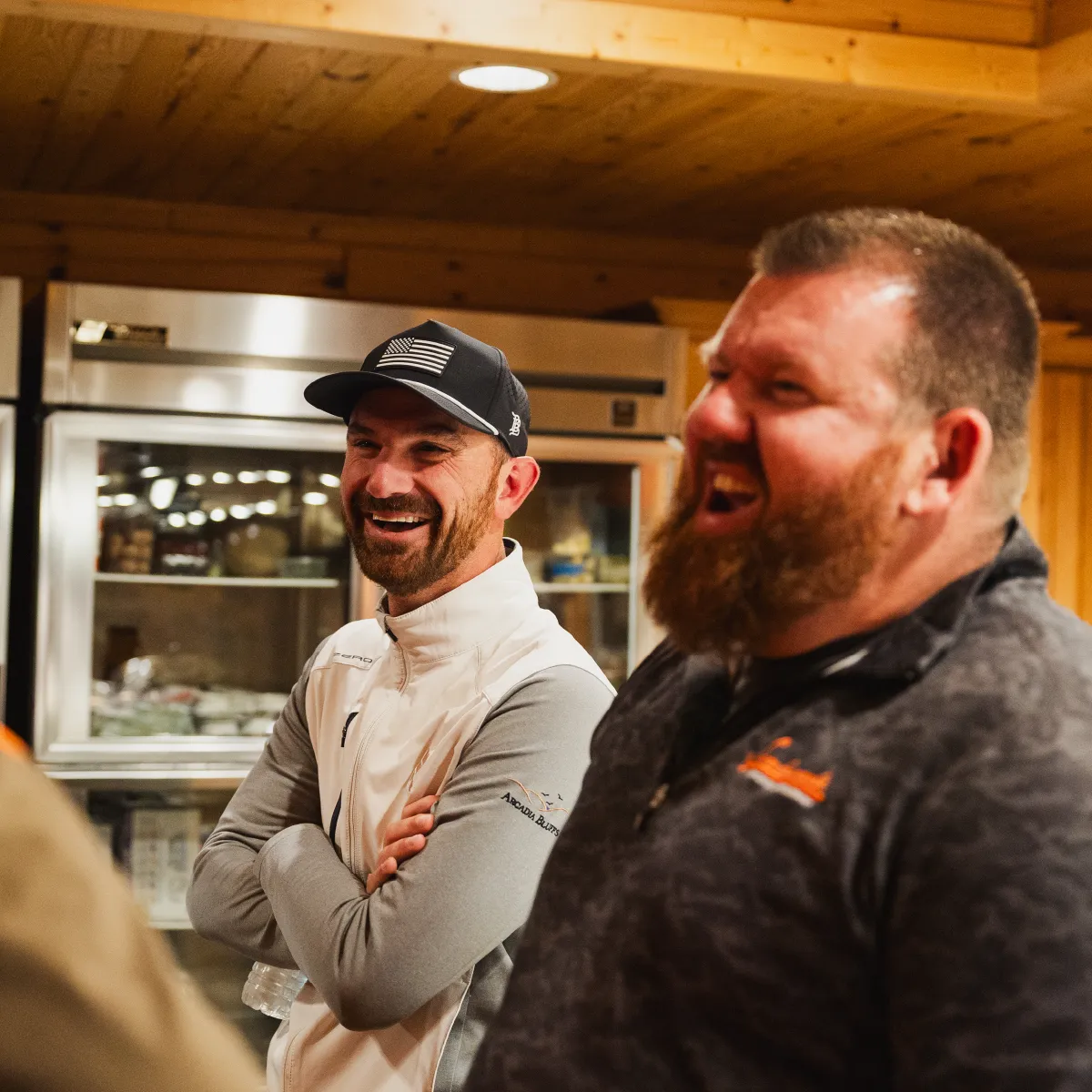 Two men laughing and chatting indoors; one wears a black cap and light jacket, the other wears a dark jacket with an orange logo.
