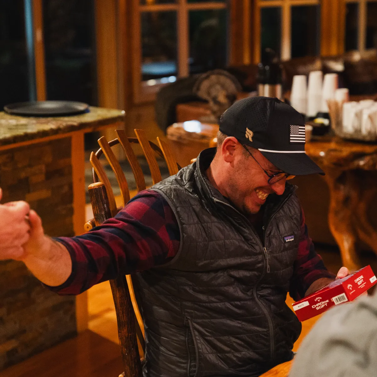 Man wearing a black cap and vest sitting at a wooden table, smiling and holding a red box labeled Chrome Soft.