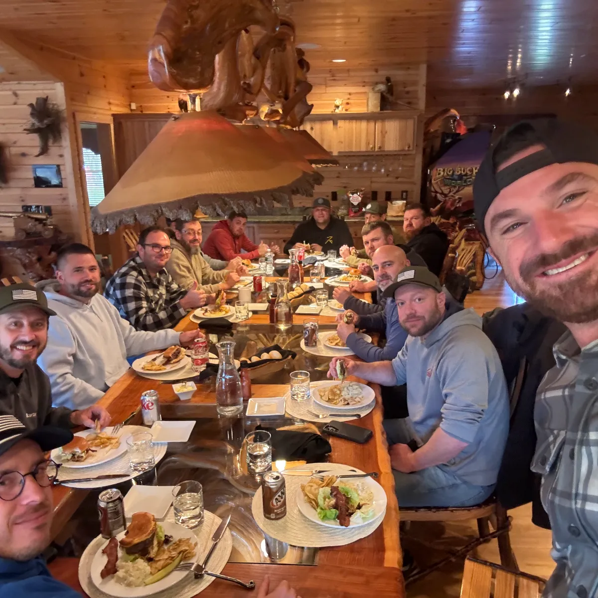 Group of men sitting around a wooden dining table in a rustic cabin, enjoying burgers and fries, with one man taking a selfie.