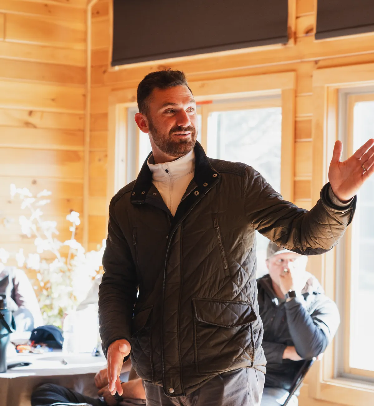 Man in a dark quilted jacket gesturing with his right hand while speaking in a wood-paneled room with large windows.