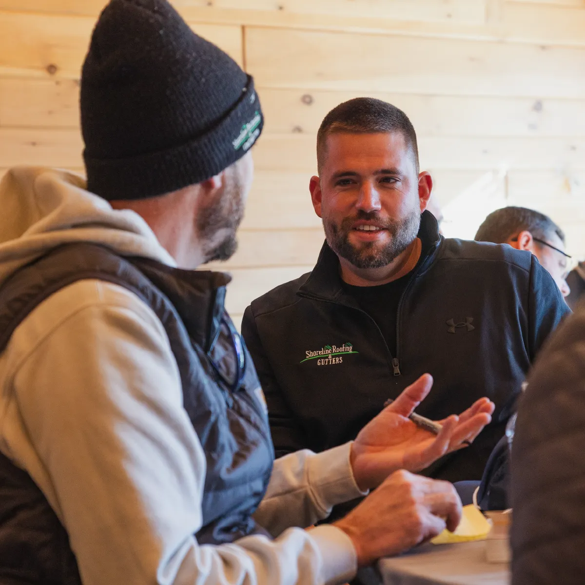 Two men in casual outdoor clothing talking indoors against a wooden paneled wall, one wearing a Shoreline Roofing & Gutters jacket.