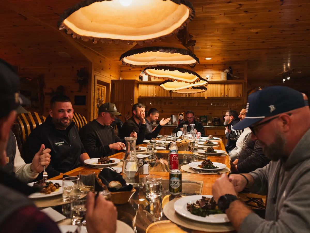 Group of people enjoying a meal together at a long wooden table under warm hanging lights in a rustic cabin.