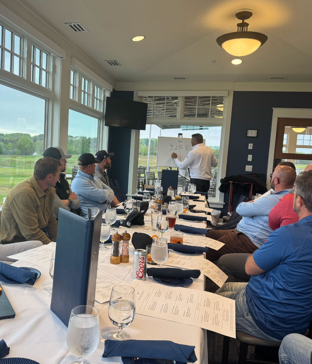Group of men seated around a long table in a bright room, attentively watching a man in a white shirt drawing on a whiteboard near large windows with a view of a golf course.