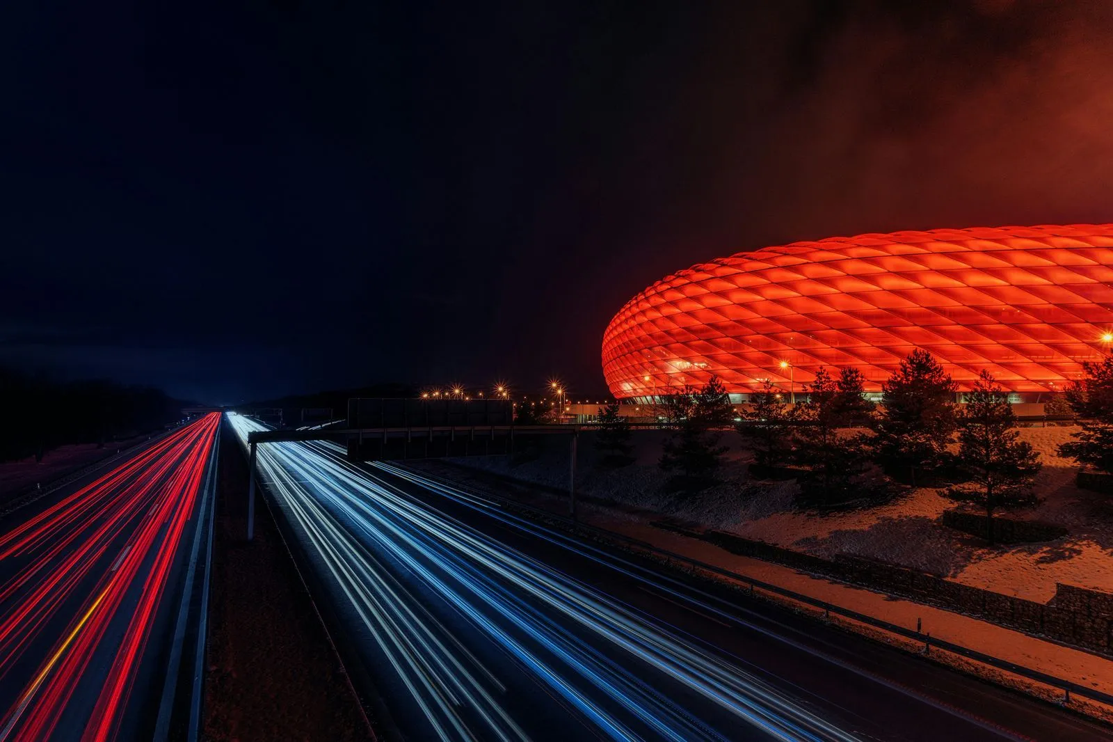 Long exposure of a highway at night with red and white light trails passing by an illuminated red stadium with a quilted facade.