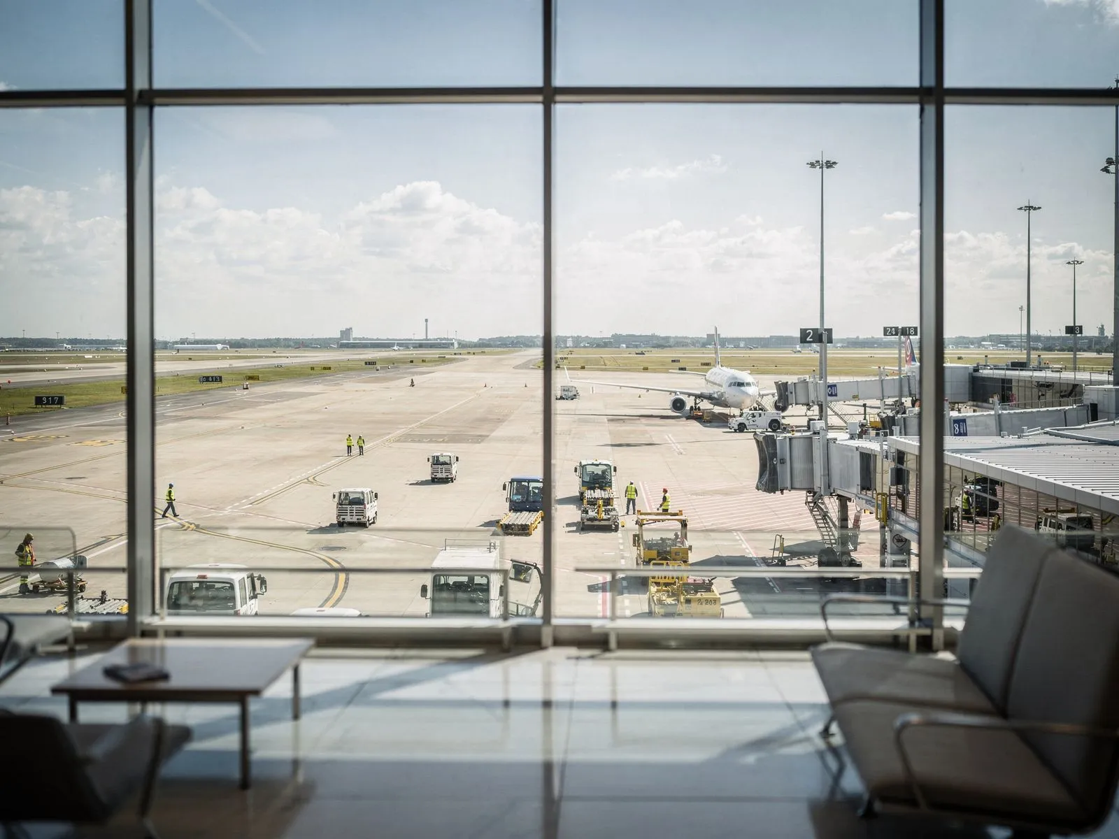 View from airport terminal seating area showing an airplane at the gate with ground crew and vehicles on the runway under a partly cloudy sky.