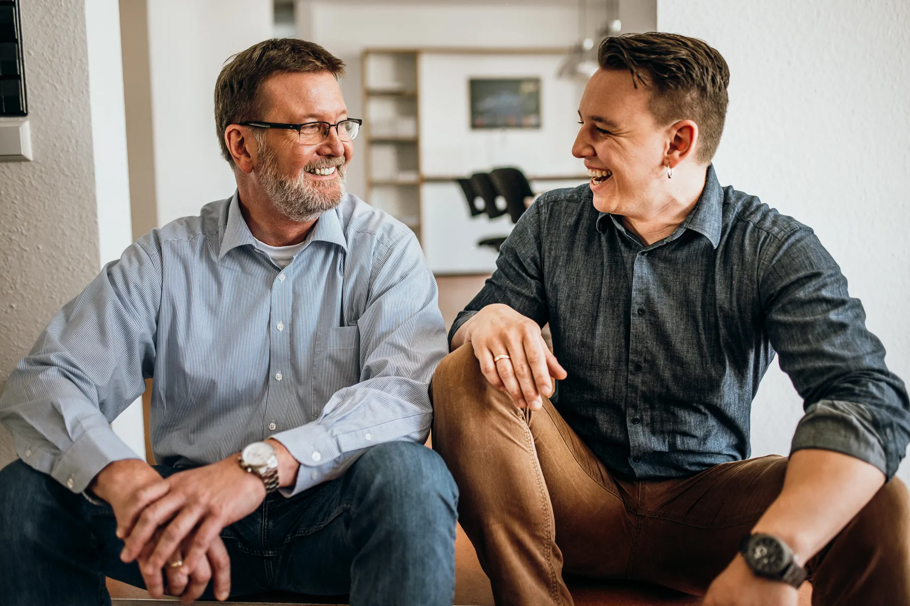 Zwei lächelnde Männer sitzen nebeneinander und schauen sich in einem Raum mit weißen Wänden an.