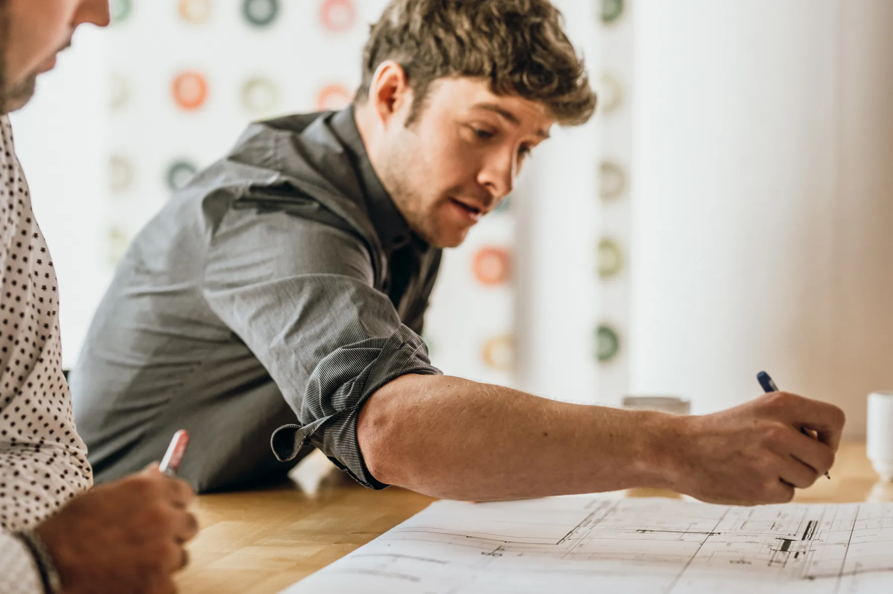Zwei Männer überprüfen zusammen einen architektonischen Plan auf einem Holztisch.