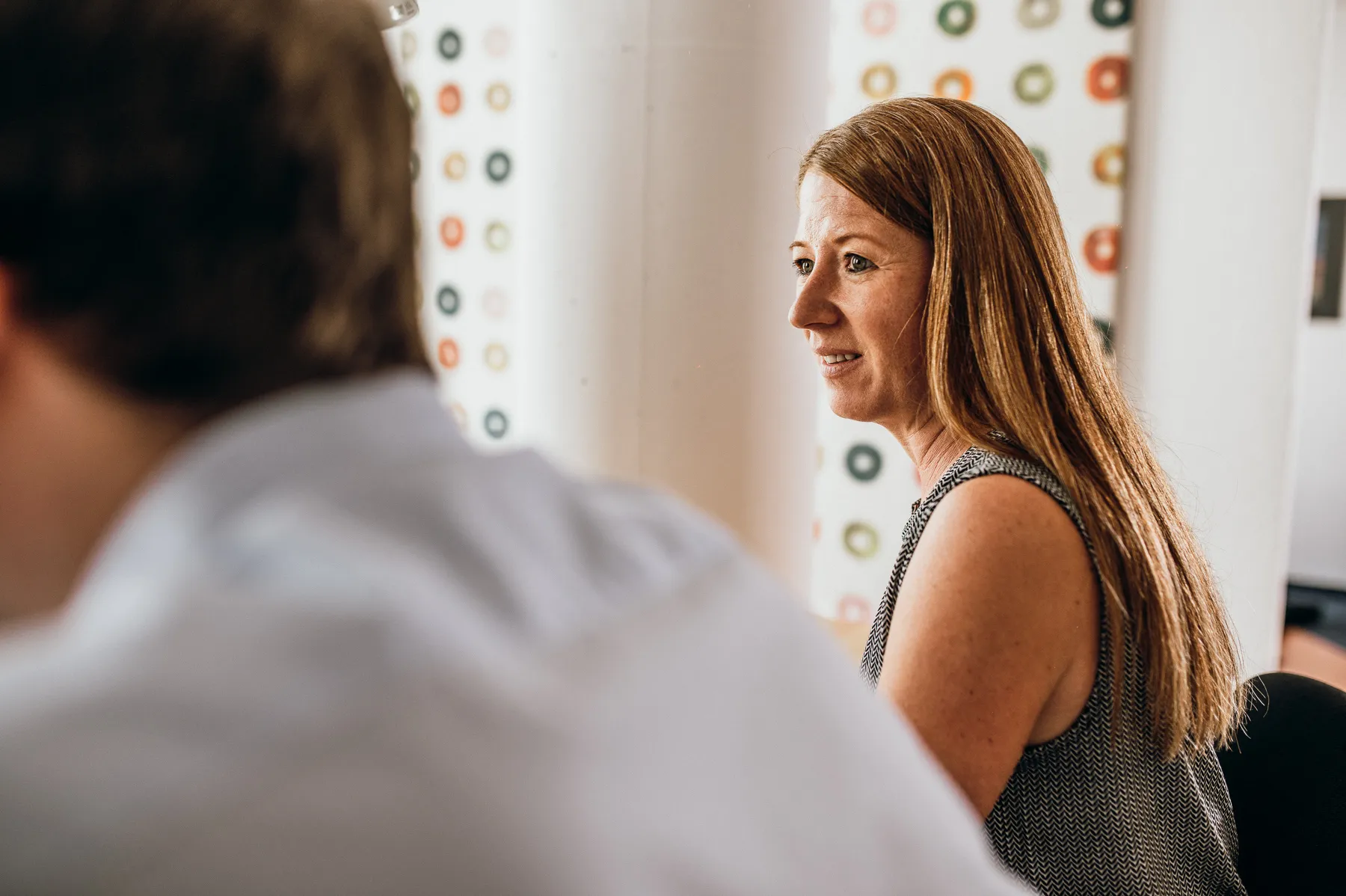 Frau mit langen roten Haaren sitzt in einem Büro und schaut aufmerksam zur Seite, im Vordergrund unscharf die Schulter einer anderen Person.
