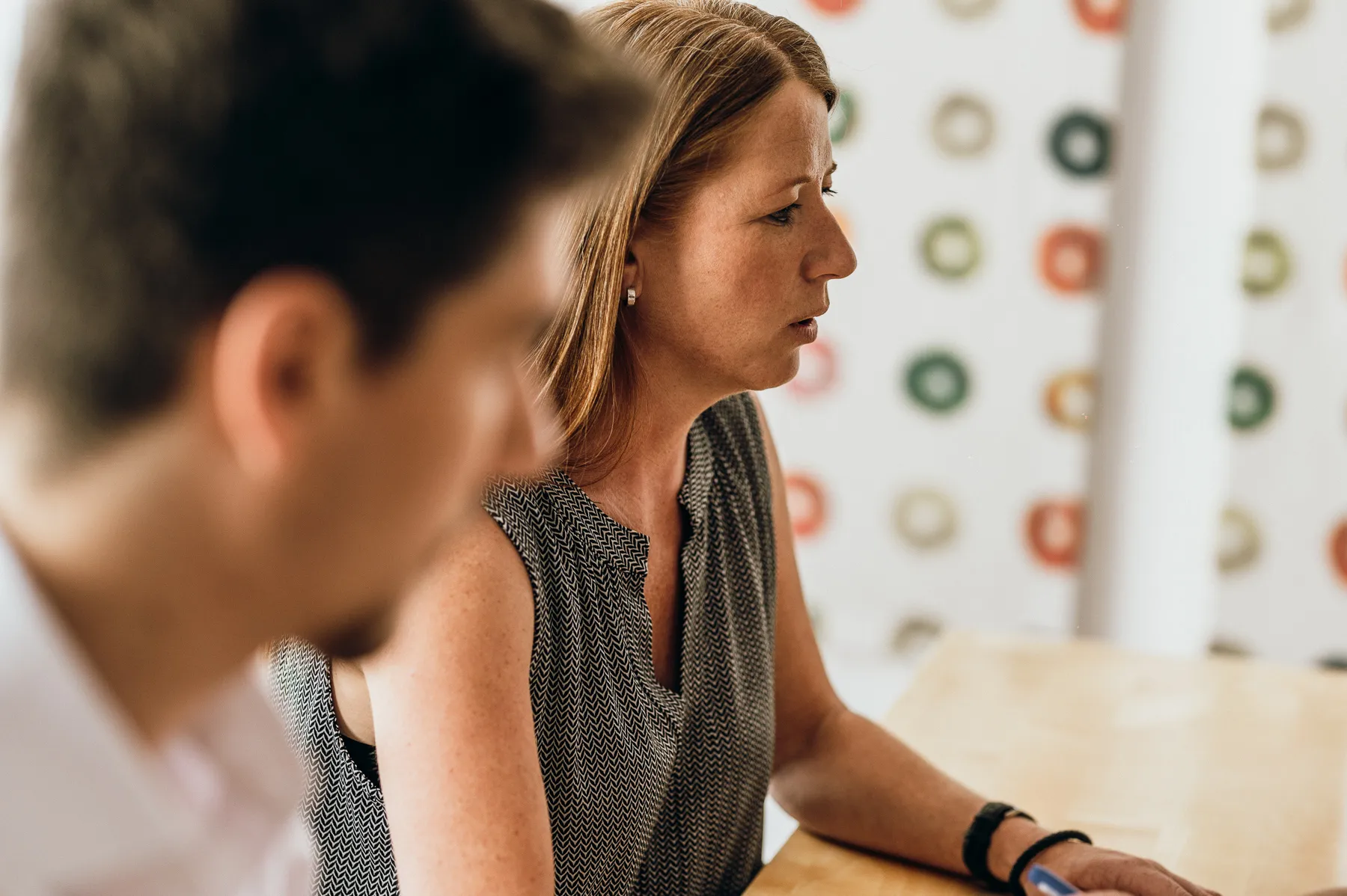 Frau mit rotem Haar und ärmellosem Top, die an einem Tisch sitzt und konzentriert wirkt, neben einem Mann im Vordergrund.