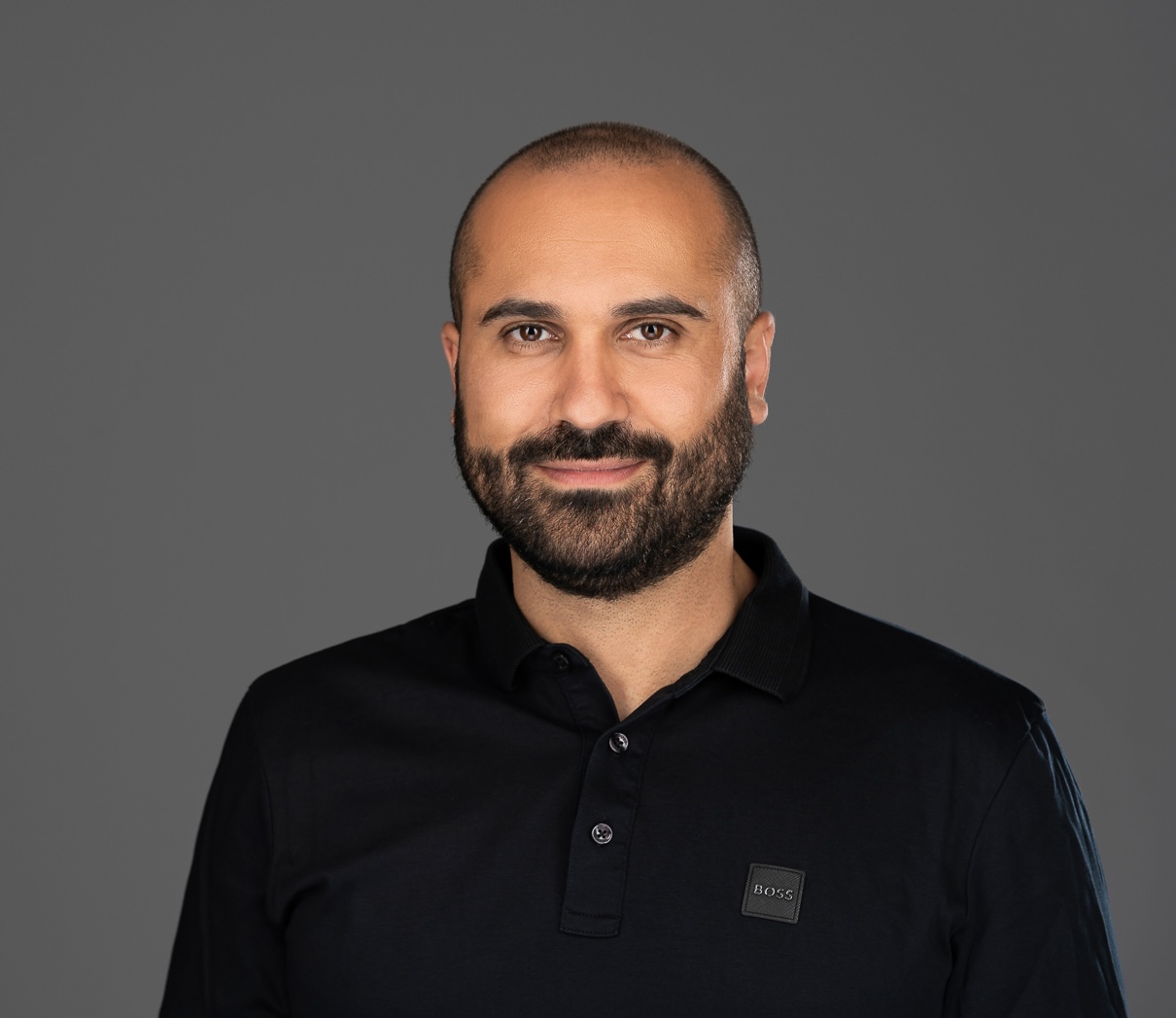 Man with a beard wearing a black Boss polo shirt smiling against a gray background.