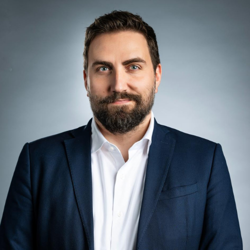 Bearded man with blue eyes wearing a white shirt and dark blazer against a gray background.