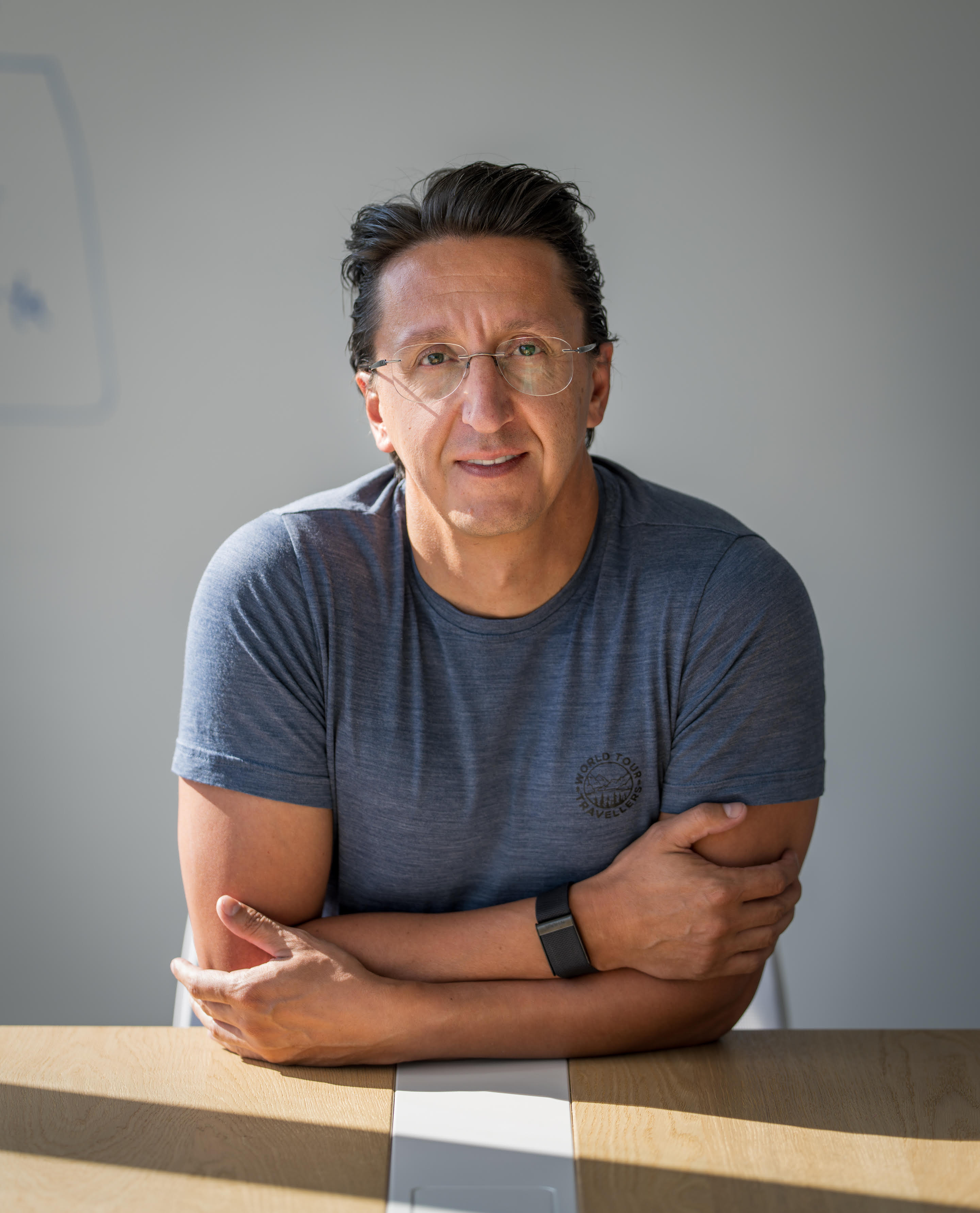 Man sits at a wooden table with arms crossed, looking forward.