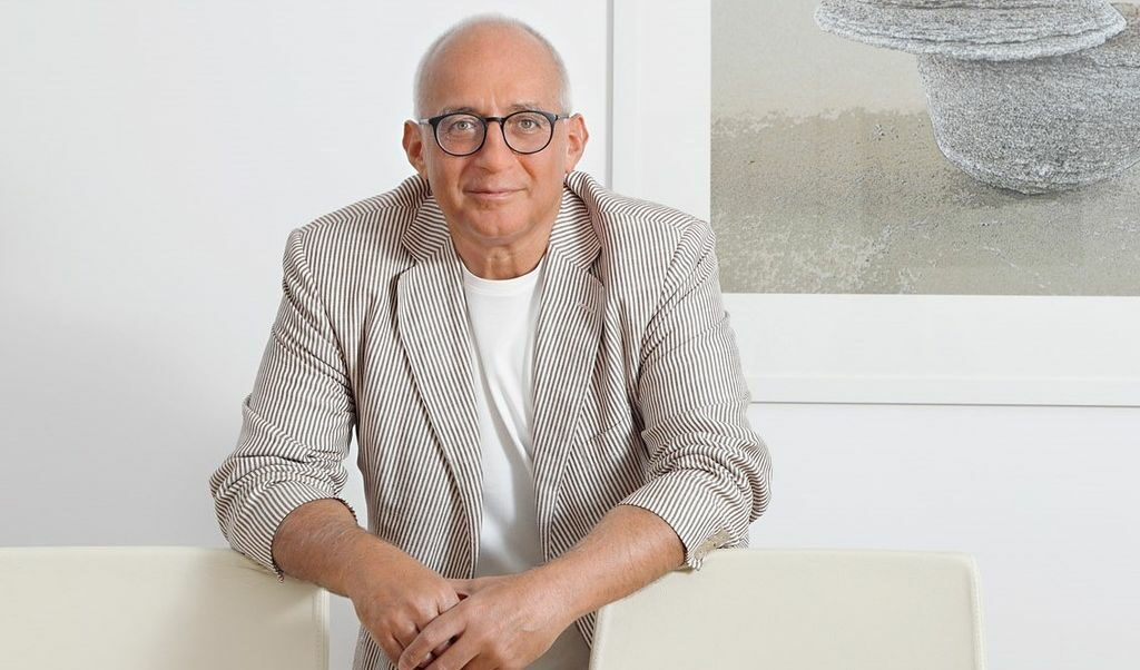 Bald man with glasses wearing a striped blazer over white shirt sitting against white wall with artwork.