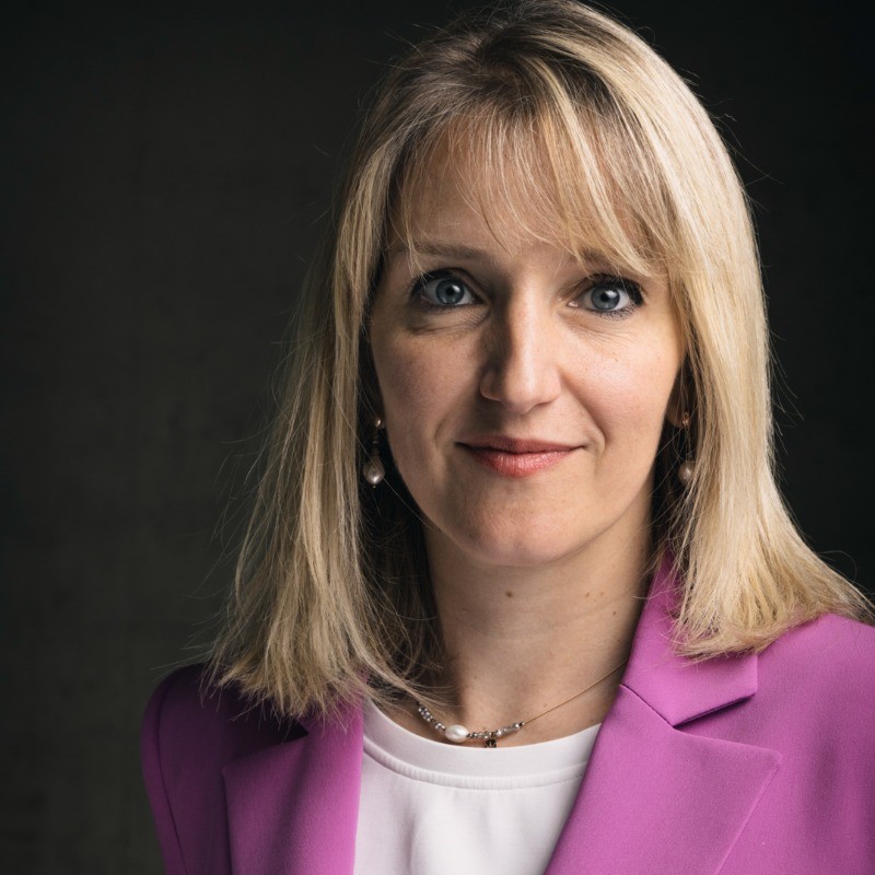 Blonde woman with blue eyes wearing a purple blazer and white shirt against a dark background.