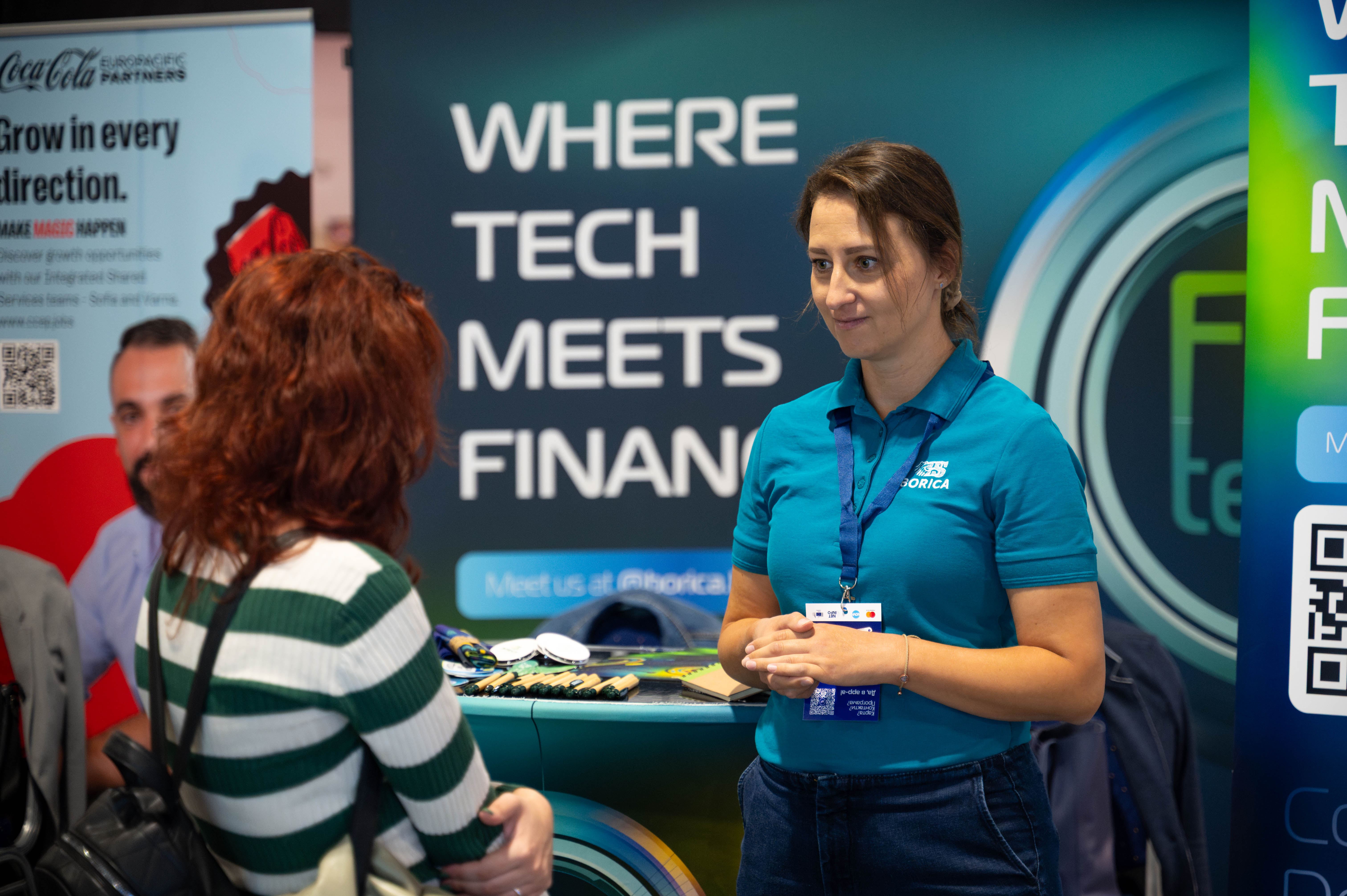 Woman in teal shirt talking to another woman in a striped sweater at a tech finance event booth.