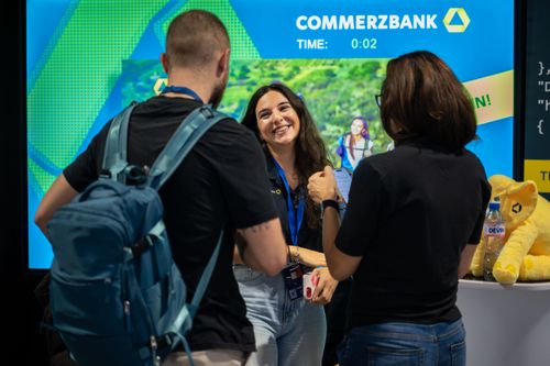 Three people talking in front of a Commerzbank display with a yellow stuffed elephant on a table.