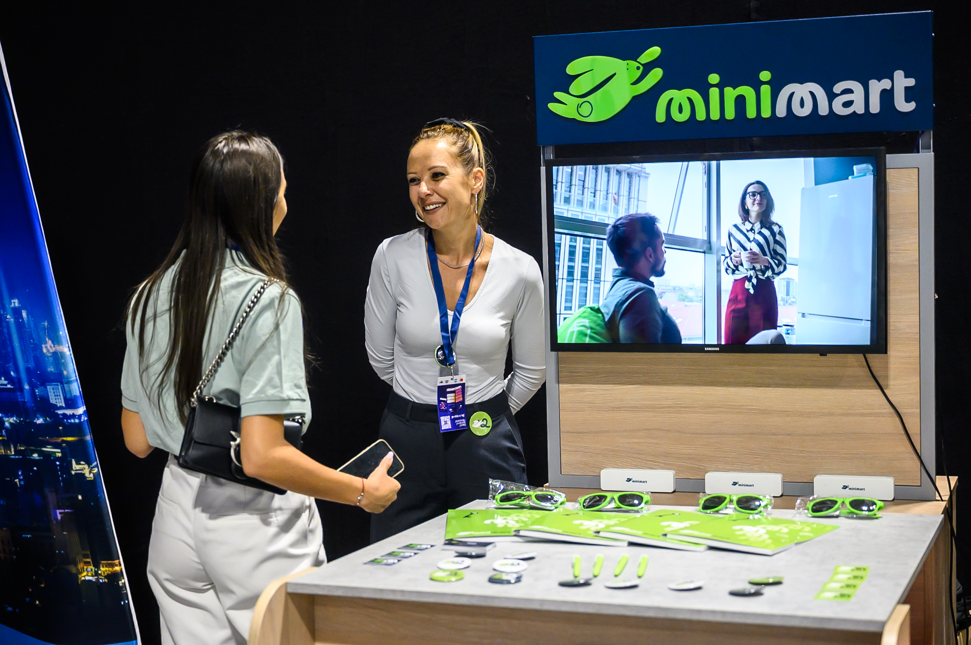 Two women talk at a minimart booth with promotional items and a screen showing two people indoors.