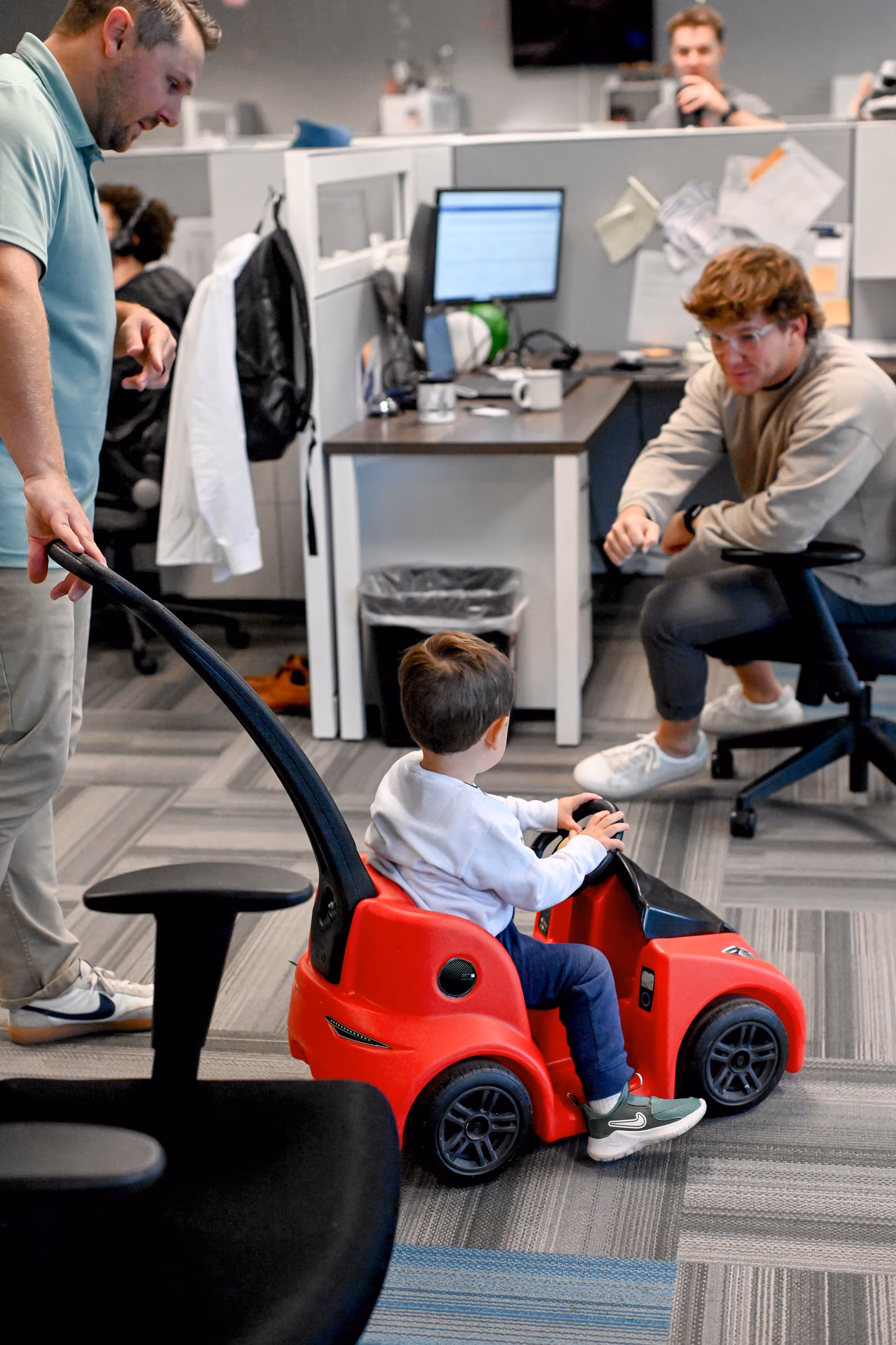 A toddler sits in a red toy car inside an office while an adult pushes the car and another adult crouches nearby engaging with the child. - Total Quality Lending Images