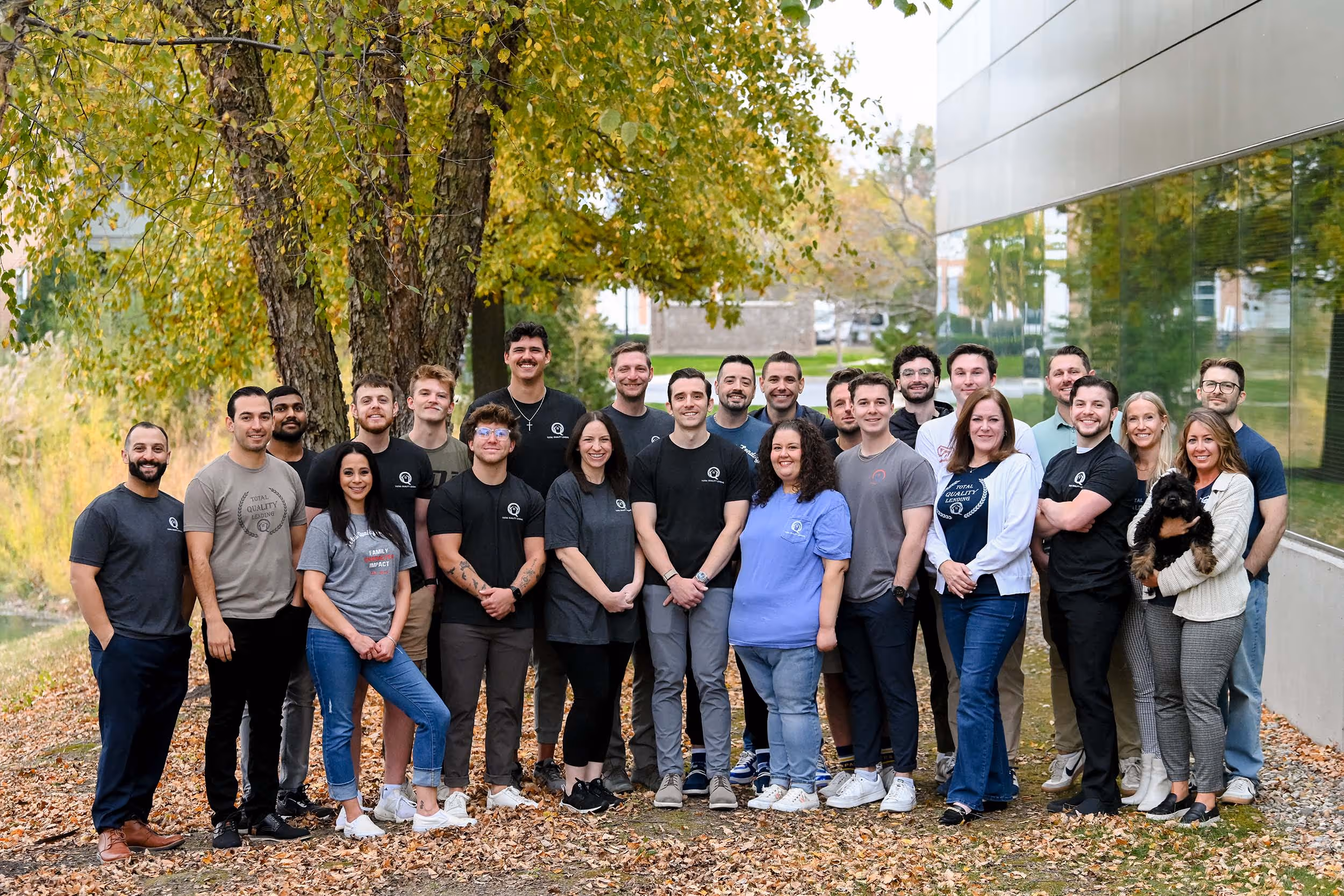 Group photo of 24 people standing together outdoors on a leaf-covered ground near trees and a modern building with glass windows, some wearing casual branded shirts. - Total Quality Lending Images