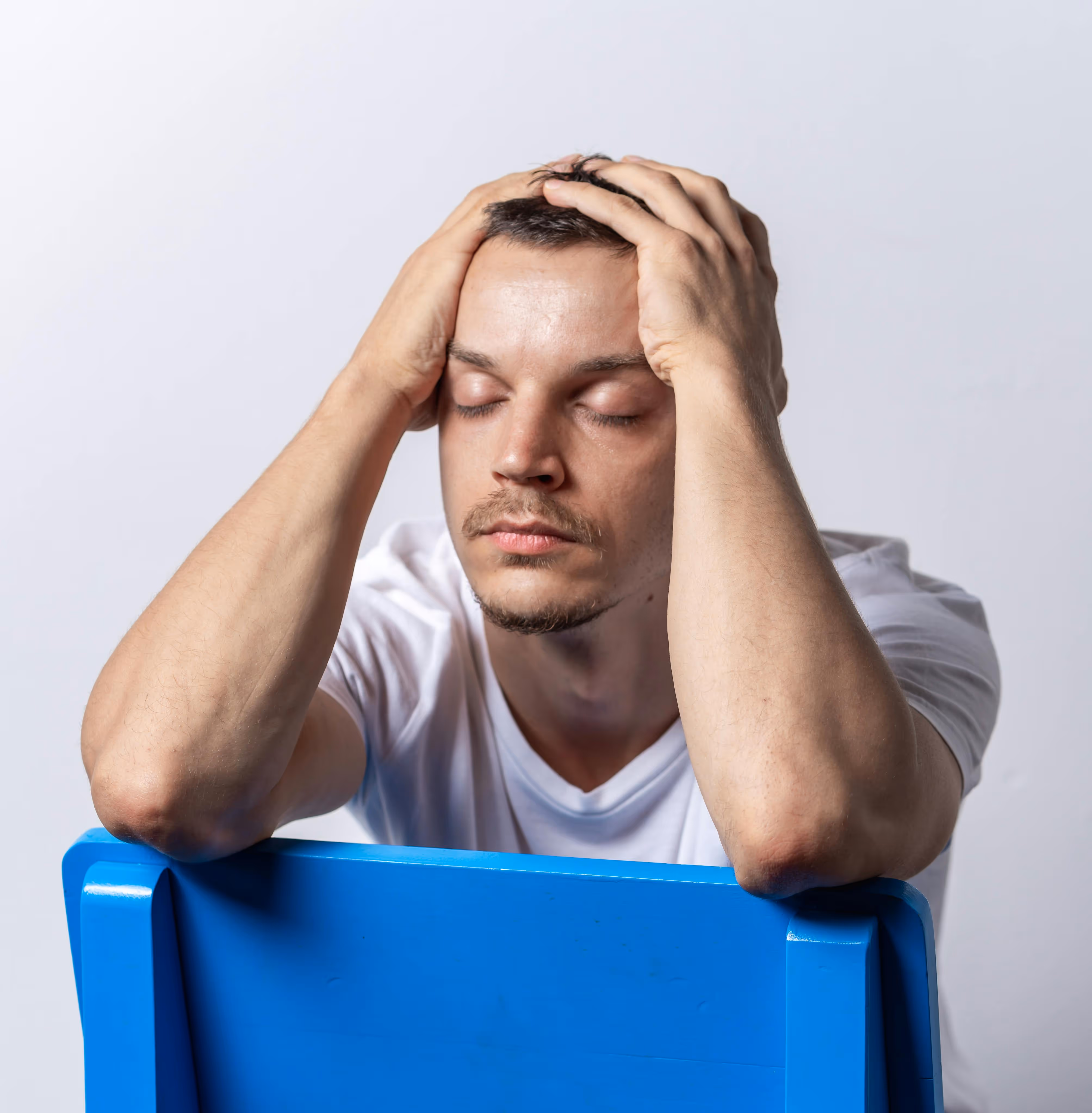 Man in a white t-shirt sitting behind a blue chair with eyes closed and hands on his head, appearing stressed. - Total Quality Lending Images