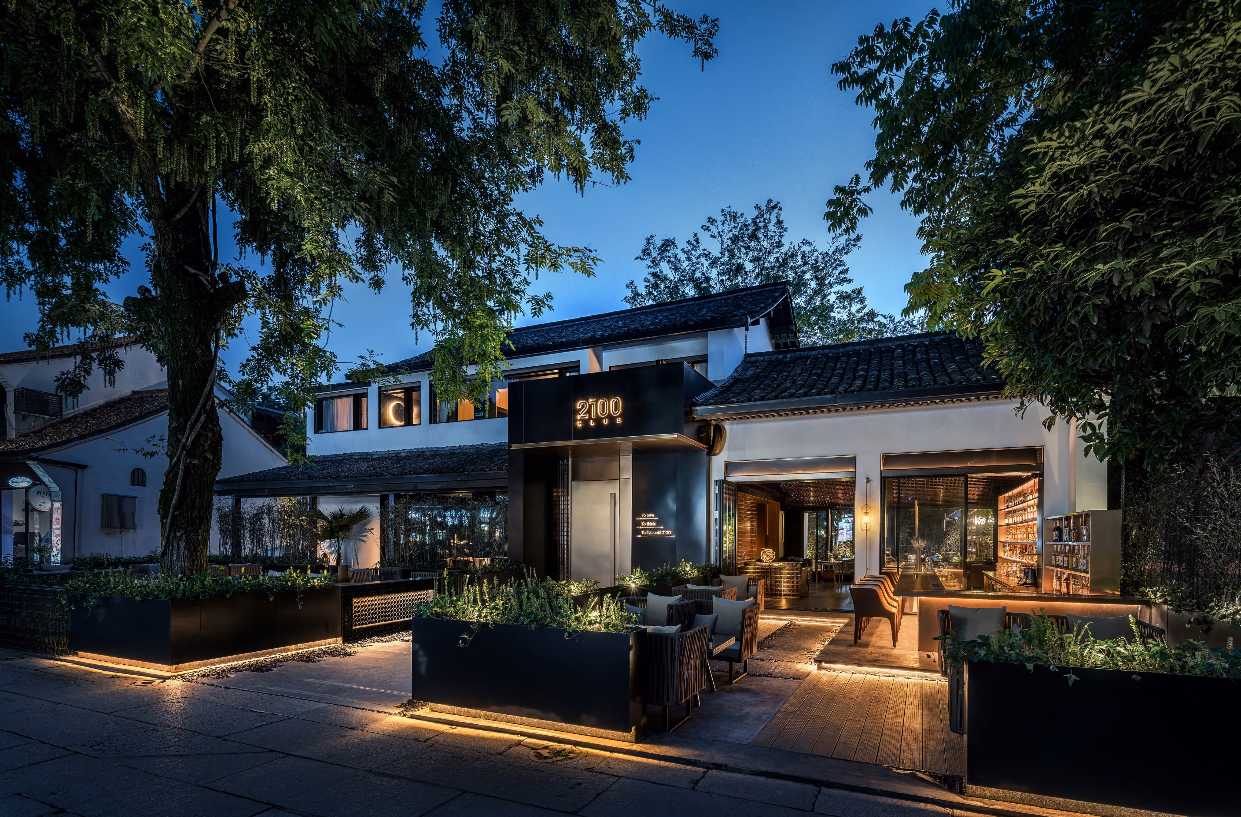 Exterior of a modern restaurant with outdoor seating and warm lighting at dusk, surrounded by trees. - Total Quality Lending Images