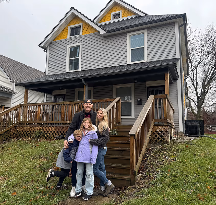 Smiling family of four standing on a grassy lawn in front of a gray two-story house with wooden porch and steps. - Total Quality Lending Images
