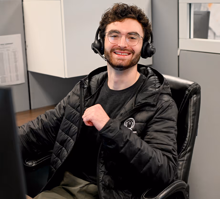 Smiling young man with glasses and headset sitting on a black office chair in a gray room. - Total Quality Lending Images