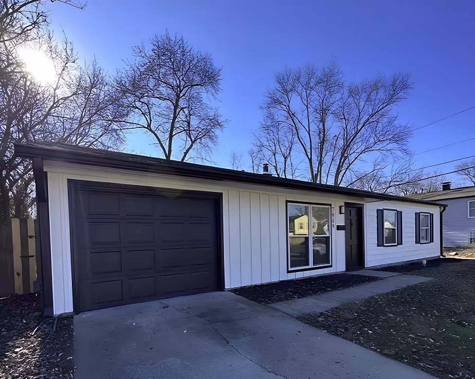 Single-story white house with black trim, a black garage door, and a driveway under a clear blue sky with leafless trees. - Total Quality Lending Images