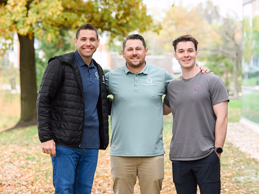 Three men standing outdoors in a park-like setting with their arms around each other, smiling at the camera on an autumn day. - Total Quality Lending Images