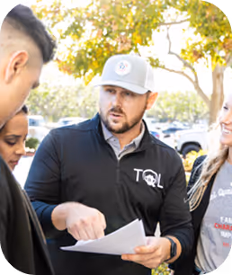 Man wearing a TQL cap and jacket explains paperwork to a small group outdoors under a tree. - Total Quality Lending Images